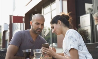 A man and woman looking at a mobile device while eating at an outdoor restaurant.