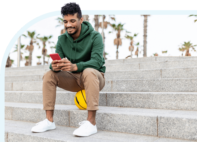 Man in green sweathshirt sitting outdoors using his mobile phone.