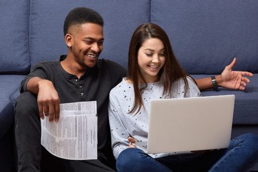 Young couple smiles as they sit by their sofa looking at a computer screen