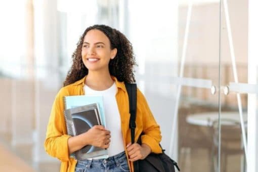 Optimistic university student with curly hair carrying notebooks and wearing a yellow shirt and backpack, standing in a modern building with glass doors.