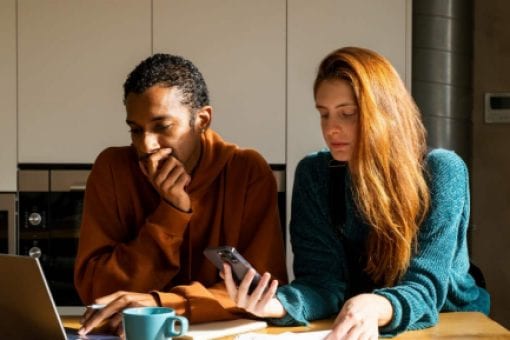 A couple sit together at a kitchen island looking thoughtfully at a laptop, mobile phone, and paperwork in front of them.