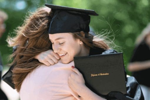 Graduate in cap and gown hugging a loved one while holding a diploma.