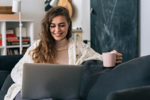 Smiling woman sitting on a couch with a laptop and holding a coffee mug in a cozy living room.