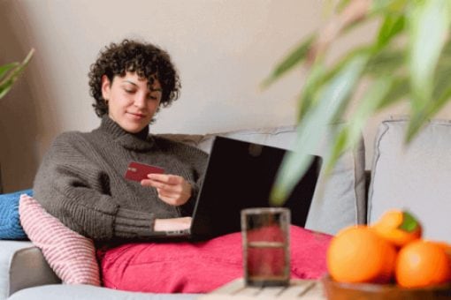 Woman sitting on a couch using a credit card to make an online purchase on a laptop at home.