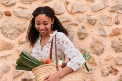 Smiling woman holding a woven basket filled with fresh vegetables, standing in front of a stone wall.