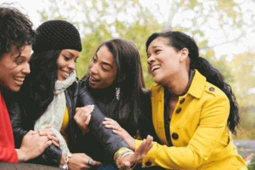 Four women sit outside in jackets. One woman holds her left hand out looking at her engagement ring as three friends embrace her and look happy.