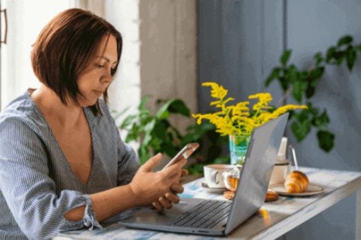 A lady sitting at a table with a laptop and phone, with breakfast items and flowers on the table.