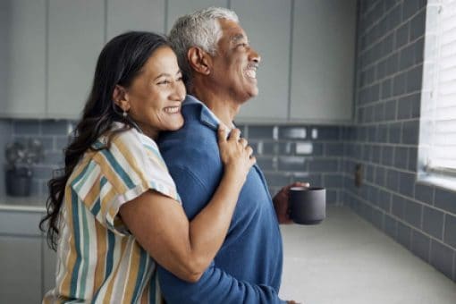 Older couple in their kitchen with the woman gently holding the man's shoulders.