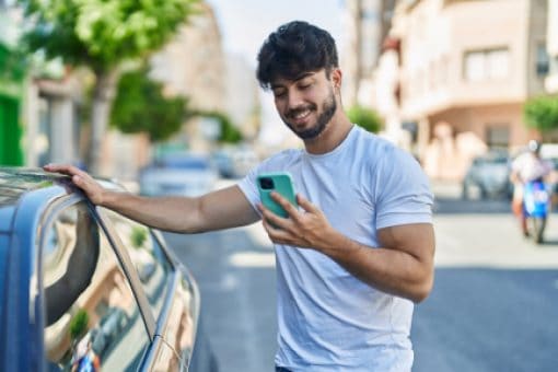 Smiling man staring at his phone leans on a car with one arm.