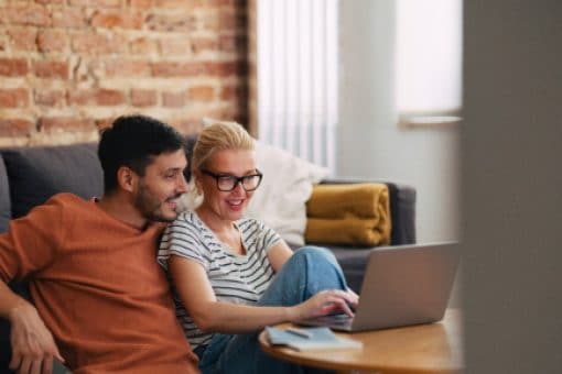 A smiling woman and man holding a cell phone at his side sit close on the floor in a cozy living room, looking together at a laptop on the coffee table.