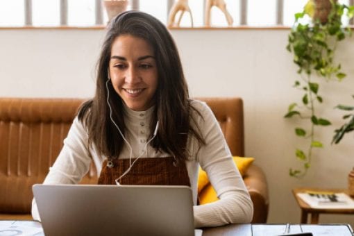 A young woman sits at a desk while excitedly working on a laptop and listening to headphones.