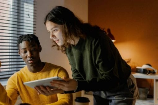 Two people collaborating at a desk, one seated with a laptop and the other standing with a tablet.