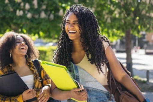 Two happy young women walk and laugh on their sunlit university campus.