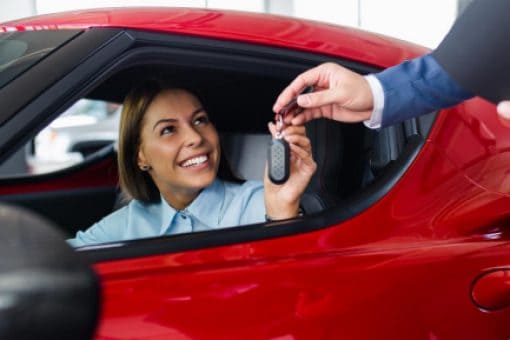 Smiling woman in a red car receiving keys after purchasing a vehicle.