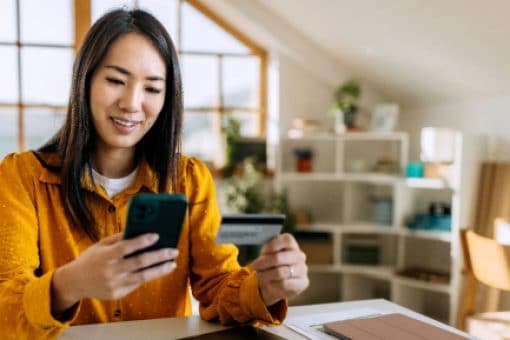 Woman sitting at a desk holding a smartphone and a credit card, smiling while making an online purchase in a well-lit home office.