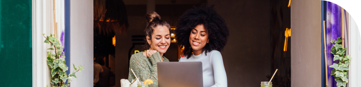 Two women sit together in an open cafe window. They both look at a laptop in front of them.