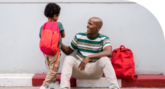 A man sits on a bench as he smiles at a young boy with a red backpack standing next to him.