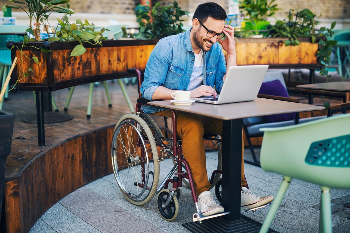 Man in a wheelchair sits at an outdoor cafe table looking at his laptop and smiling.